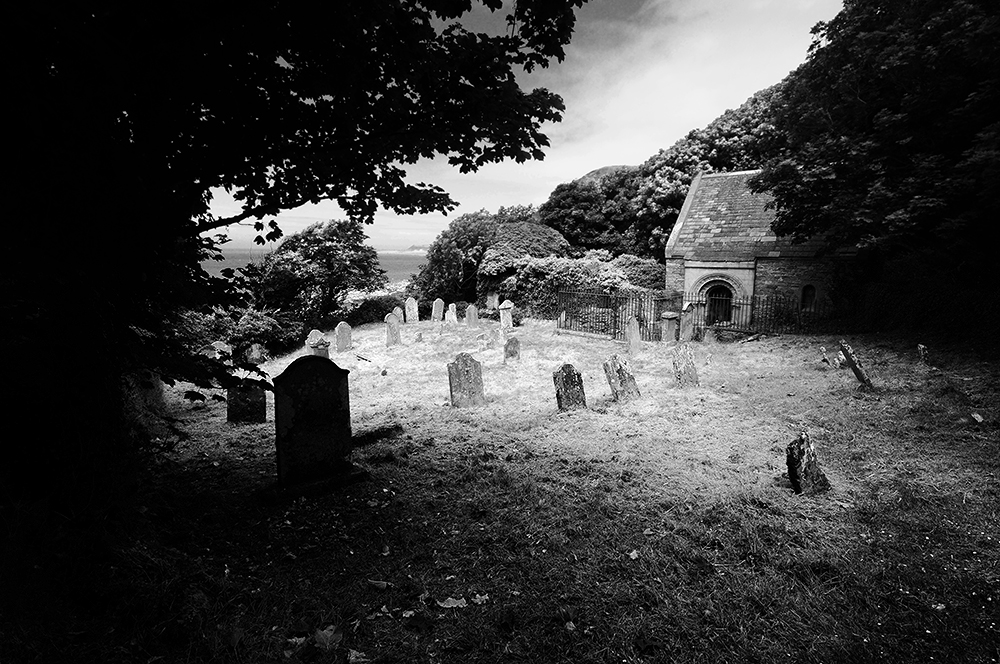 Black and white photo of a ruined church and graveyard, with old, leaning tombstones.