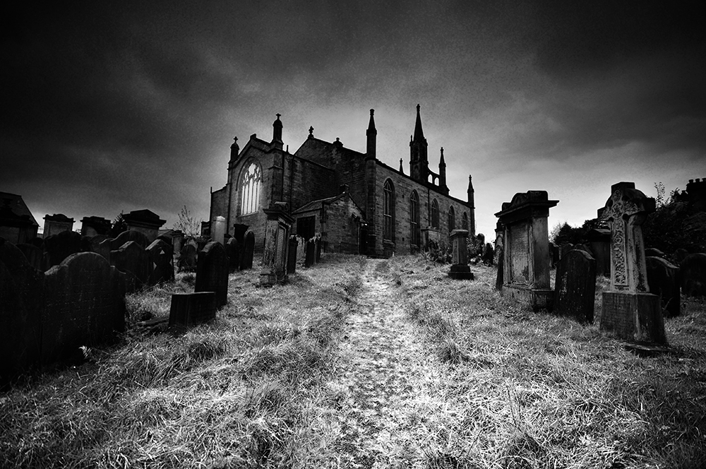 Black and white photo of a gothic church and graveyard  on a hill, with dark clouds above.
