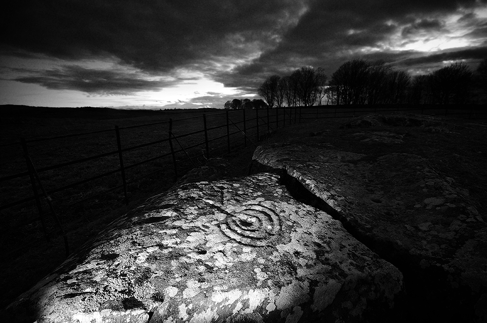 Black and white image of a rock outcrop bearing cup and ring markings. The sun has set behind the trees on the near horizon and a dark gloming sky is overhead.